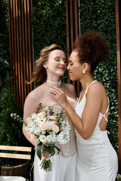 Две невесты обнимаются в день своей свадьбы, окруженные пышной зеленью.