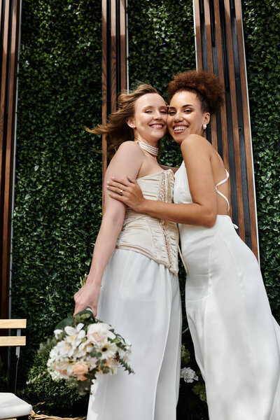 Две невесты обнимаются в день своей свадьбы, радостный момент, запечатленный перед пышной зеленой стеной.