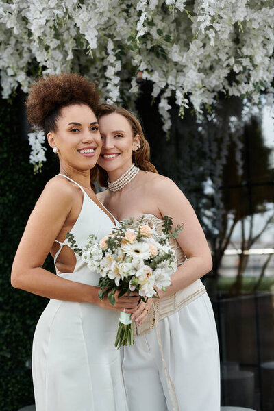 Две невесты стоят рядом, одна держит букет белых цветов, во время их свадебной церемонии.