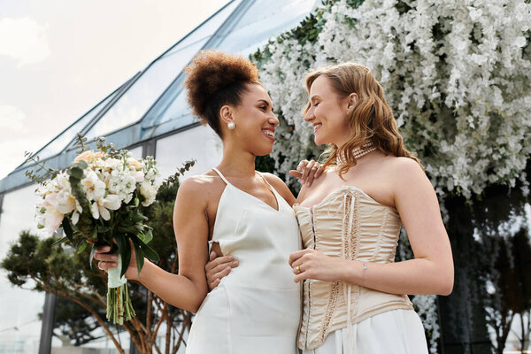 A lesbian couple smiles at each other during their wedding ceremony, surrounded by white flowers.