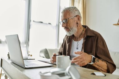 Dövmeleri ve gri saçı olan olgun bir eşcinsel ev ofisinden uzaktan çalışıyor..