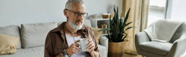 Gri saçlı ve dövmeli olgun bir adam evde çalışırken bir fincan kahveden zevk alır..