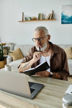 A mature gay man with tattoos and gray hair sits at a desk in his home office, working remotely while holding a notebook and pen. clipart