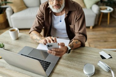 Gri saçlı ve dövmeli olgun bir eşcinsel, akıllı telefon ve dizüstü bilgisayar kullanarak evden uzakta çalışır..