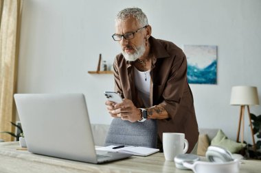 Dövmeleri ve gri saçı olan olgun bir eşcinsel evde çalışırken telefonunu kontrol ediyor..