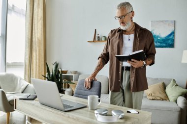 A tattooed older man with gray hair works from home, standing by his desk with a notebook, focused on his work. clipart