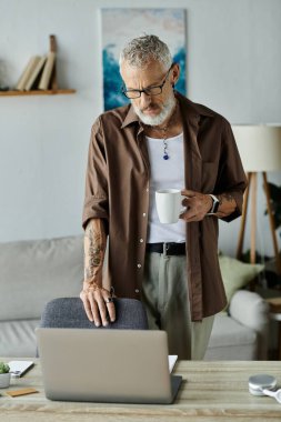 Evde uzaktan çalışan, dövmeli ve gri saçlı, olgun bir gay, dizüstü bilgisayarı üzerinde çalışırken bir fincan kahveden zevk alır..