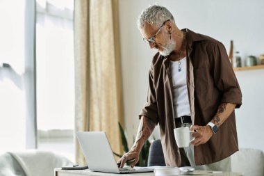 Olgun, dövmeli, gri saçlı bir adam elinde bir fincan kahve tutarken dizüstü bilgisayarında çalışıyor..