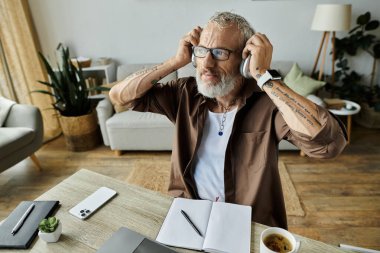 Dövmeleri ve gri saçları olan olgun bir eşcinsel kulaklık takarak evde çalışır..