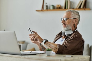 Dövmeleri ve gri saçı olan olgun bir eşcinsel, akıllı telefonu ve dizüstü bilgisayarıyla evden uzakta çalışır..