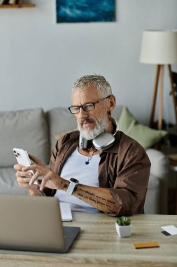 Dövmeleri ve gri saçı olan olgun bir adam evinden uzakta çalışıyor. Kulaklık takarken akıllı telefon ve laptop kullanıyor..