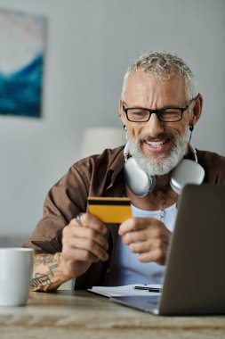 Gri saçlı ve dövmeli olgun bir eşcinsel elinde kredi kartıyla gülümseyip laptopuna bakıyor ve evden çalışıyor..