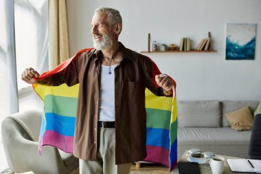 Dövmeleri ve gri saçı olan olgun bir gay evde bir gökkuşağı gururu bayrağı tutuyor, gülümsüyor..