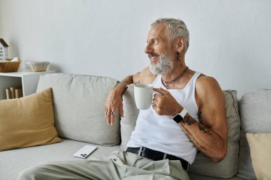 Dövmeli, gri sakallı, olgun bir gay evdeki kanepesinde bir kupa kahveyle rahatlar ve düşünceli bir şekilde kamera önünde görünmez olur..