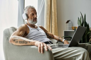 A mature gay man with tattoos and a grey beard rests on a couch, wearing headphones and looking at a laptop. clipart