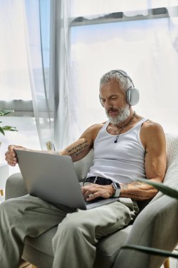 Dövmeli, gri sakallı bir adam dizüstü bilgisayar kullanırken koltuğunda dinleniyor..