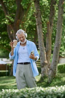 Dövmeli bir adam, gri sakallı, güneşli bir parkta kahve eşliğinde müzik dinlemekten hoşlanıyor..
