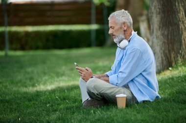 A mature gay man with tattoos and a gray beard sits on the grass in a park, scrolling on his phone. clipart