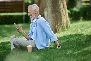 A mature gay man with tattoos and a grey beard smiles while using his phone in a park. clipart