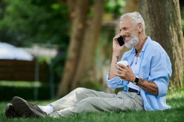 A mature gay man with tattoos and a grey beard sits on the grass, smiling while talking on his phone. clipart