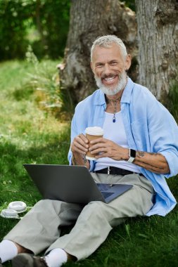 Gri sakallı ve dövmeli olgun bir adam parkta çimlerin üzerinde oturur, kahve fincanı tutarken gülümser ve dizüstü bilgisayar kullanır..