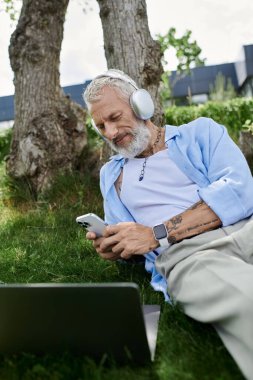 Dövmeli ve gri sakallı olgun bir adam çimlerin üzerinde oturur, kulaklıkla müzik dinler ve telefonuna bakar..