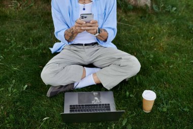 Dövmeli ve gri sakallı olgun bir adam dışarıdaki çimlerin üzerinde telefonuyla oturuyor..