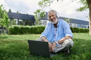 Dövmeli, gri sakallı, kulaklık takmış ve kameraya gülümseyen olgun bir eşcinsel..