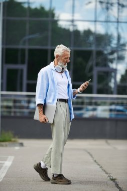 Gri sakallı ve dövmeli olgun bir adam elinde dizüstü bilgisayarla şehir caddesinde yürür ve telefonunu kontrol eder..
