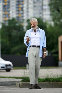 Gri sakallı, dövmeli yaşlı bir adam şehir caddesinde duruyor, telefonunu kontrol ediyor..
