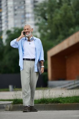 Dövmeli, olgun, gri sakallı eşcinsel bir adam şehirde yürürken cep telefonuyla konuşurken gülümsüyor..