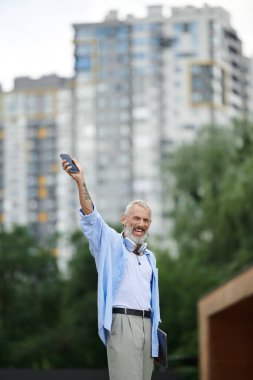 Dövmeli ve gri sakallı olgun bir eşcinsel, şehir parkında beklerken neşeli bir şekilde gülümsüyor..
