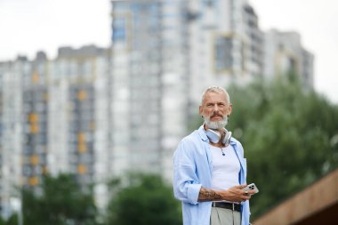 A mature man with tattoos and a grey beard standing in an urban setting. clipart