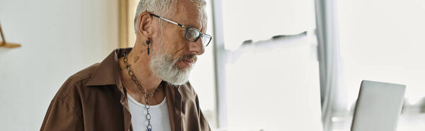 Взрослый гей с татуировками и седыми волосами работает дистанционно на своем ноутбуке дома.