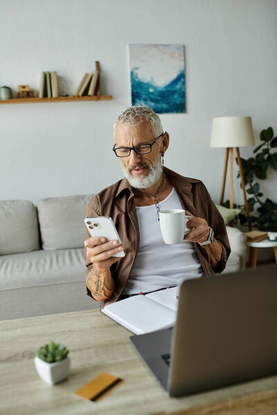 A tattooed, mature gay man with grey hair works remotely from home while holding a coffee mug and looking at his phone.