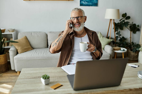 A mature gay man with tattoos and grey hair smiles while talking on his phone and sipping coffee during a remote workday.