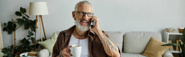 A mature, tattooed gay man with grey hair smiles while talking on the phone in his home.