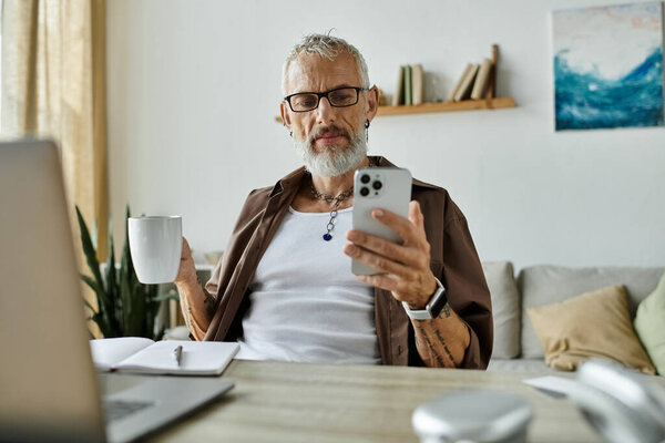 A mature gay man with tattoos and grey hair works remotely from home, holding a coffee mug and looking at his phone.