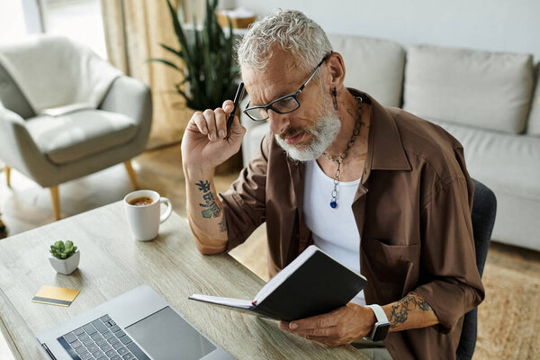 A mature gay man with grey hair and tattoos works remotely from home, sitting at a desk with a laptop and a notebook.