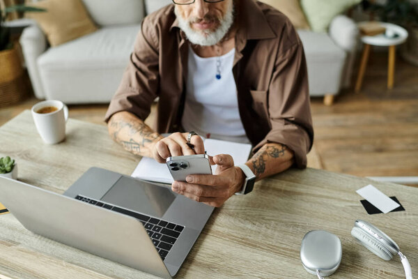 A mature gay man with grey hair and tattoos works remotely from home, using a smartphone and laptop.