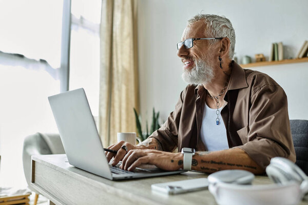 A mature gay man with tattoos and grey hair works remotely from home, smiling and typing on a laptop.