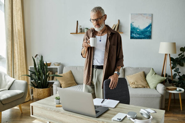 A mature gay man with tattoos and gray hair works from home, taking a break to enjoy a cup of coffee.