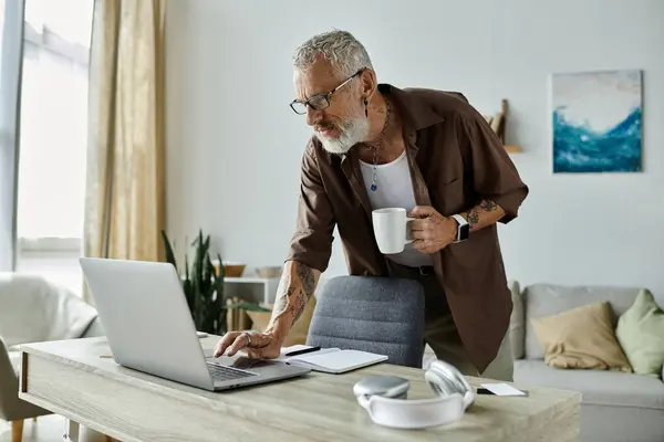 stock image A mature, tattooed gay man works remotely from home, standing at his desk with a laptop and a cup of coffee.