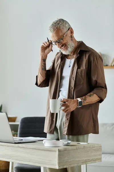 stock image A mature gay man with tattoos and grey hair enjoys a cup of coffee while working remotely from home.