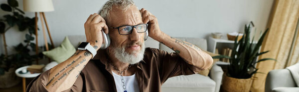 A mature gay man with tattoos and grey hair, wearing glasses and headphones, works remotely from home.
