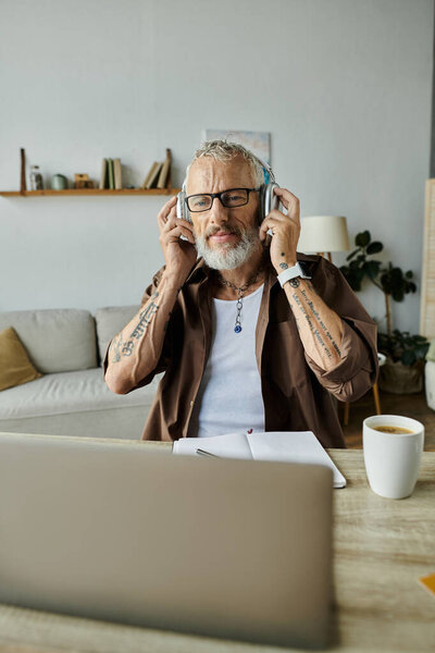 A mature gay man with tattoos and grey hair works remotely from home while wearing headphones.