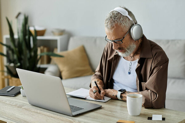 A mature gay man with grey hair and tattoos works remotely from home, taking notes in a notebook while wearing headphones.