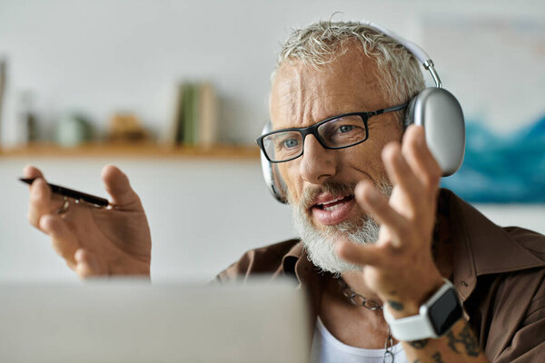 A mature gay man with grey hair and tattoos works remotely from home while wearing headphones and glasses.