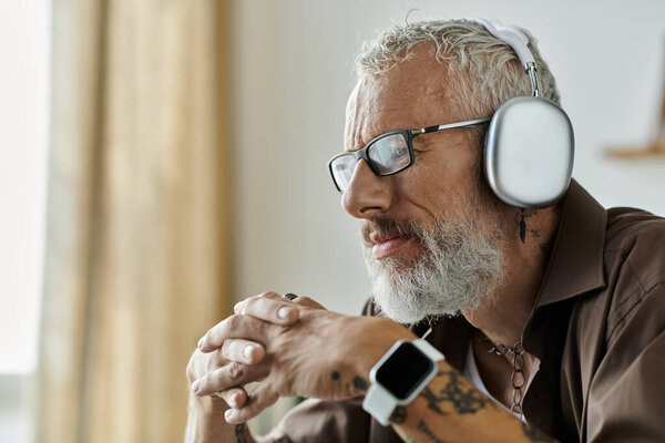 A mature gay man with tattoos and grey hair is working remotely from home. He is wearing glasses and headphones.