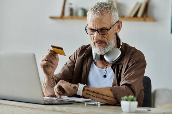 A mature man with grey hair and tattoos works remotely from home, holding a credit card and looking at a laptop.
